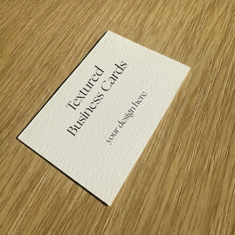 May include: A white textured business card with the text "Textured Business Cards" and "your design here" printed in black. The card is angled on a light brown wooden surface.