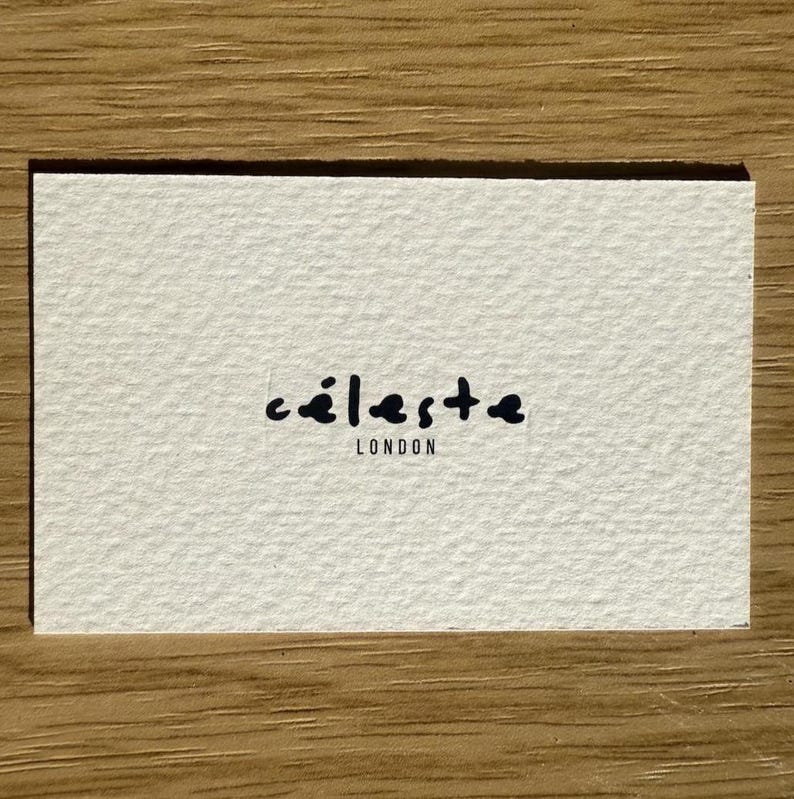 May include: A rectangular, off-white business card with the word "c&eacute;leste" in a handwritten-style font, followed by "LONDON" in a smaller font. The card has a textured surface.