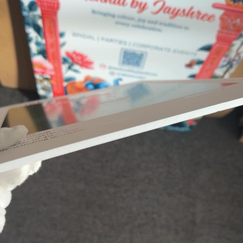 May include: A white rectangular sign with text and a floral background. The sign is held at an angle, revealing its edge. The background features red pillars and floral patterns. The sign appears to be for a business offering event services.