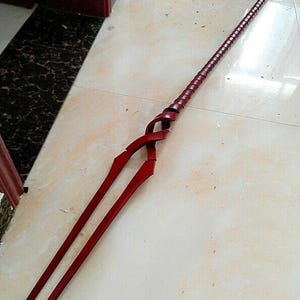 May include: A long, red spear with a spiraled handle and a forked tip. The spear is made of a dark red material and appears to be a prop or decorative item. The spear is lying on a light-colored tiled surface.