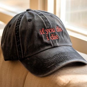 May include: A black, distressed baseball cap with the embroidered phrase "if you die i die" in red thread. The cap is sitting on a beige upholstered surface near a window.