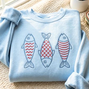 May include: A light blue crewneck sweatshirt featuring an embroidered design of three fish arranged side by side. Each fish is outlined in blue thread with distinct red patterns on their bodies, including wavy lines, scales, and horizontal stripes.