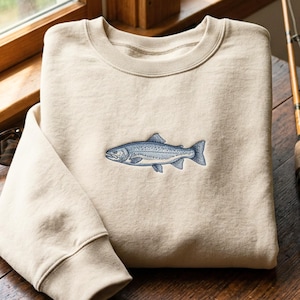May include: A beige sweatshirt featuring a blue fish embroidery. The garment is displayed on a wooden surface, with a fishing rod and sunglasses visible. The fish design is detailed. The sweatshirt is a casual item.