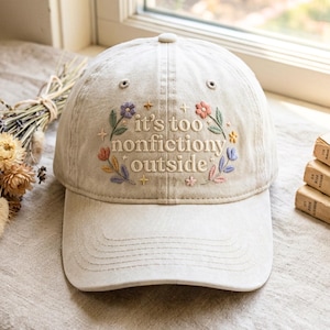 May include: A beige baseball cap featuring embroidered text that reads "it's too nonfictiony outside" surrounded by small colourful flowers and star shapes. The cap has a curved brim and stitched eyelets on the crown for ventilation.