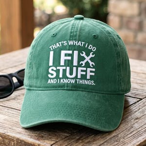 May include: A green baseball cap with the text "THAT'S WHAT I DO, I FIX STUFF AND I KNOW THINGS" in white. The cap is sitting on a wooden table next to a pair of black sunglasses. The cap has a curved brim.