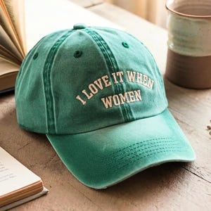 May include: A green baseball cap with a distressed finish featuring the embroidered text I LOVE IT WHEN WOMEN in a cream-colored font. The cap has a structured crown, a curved brim with visible stitching, and eyelets for ventilation on the top panels.