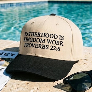 May include: A tan and black baseball cap with the text "FATHERHOOD IS KINGDOM WORK PROVERBS 22:6" embroidered on the front. The cap has a curved brim and a black button on top. Sunglasses and a magazine are next to the cap.