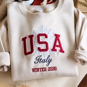 May include: Cream-colored sweatshirt with the word "USA" in red, outlined in white, above "Italy" and "Winter 2026" in navy blue. A snowflake design is behind the word "USA". The sweatshirt is folded neatly.