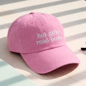 May include: A pink baseball cap with the embroidered text "hot girls read books" in white. The cap is made of a soft fabric and has a curved brim. A pair of eyeglasses rests on a wooden surface.