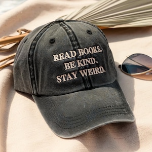 May include: A dark gray baseball cap with the embroidered text "READ BOOKS. BE KIND. STAY WEIRD." The cap is sitting on a light-colored surface, with sunglasses resting nearby. The hat has a curved brim and a classic six-panel design.