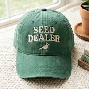 May include: A forest green baseball cap featuring the embroidered text SEED DEALER in a cream-colored font, positioned above a small, simple embroidered graphic of a bird facing right, centered on the front panel of the six-panel crown.