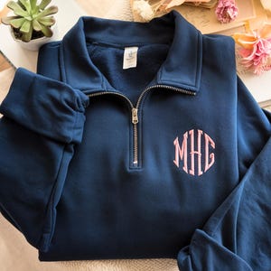 May include: A navy blue quarter-zip sweatshirt with a pink monogram. The sweatshirt features a collared neckline and a gold zipper. The monogram is embroidered on the left chest. The sleeves are cuffed. The sweatshirt is displayed on a white surface.