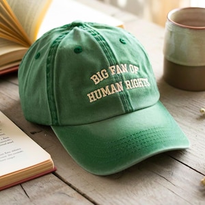 May include: A green baseball cap with the embroidered text "BIG FAN OF HUMAN RIGHTS" in white. The cap is sitting on a wooden surface next to an open book and a mug.