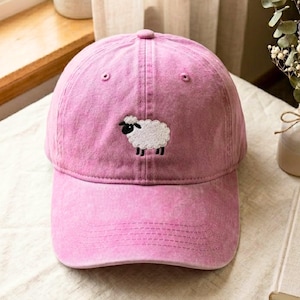 May include: A pink baseball cap with a distressed, washed-out finish. The front panel features a small, centered embroidery of a white sheep with a black face and black legs. The cap has a curved brim and a structured crown with stitched eyelets.