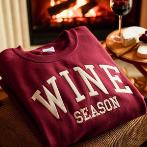 May include: A burgundy sweatshirt with the words "WINE SEASON" in cream-colored, block letters. The sweatshirt is folded and resting on a woven chair. A glass of red wine, cheese, and grapes are on a small table.