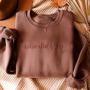 Peut inclure: Un sweat-shirt marron &agrave; col rond avec le texte brod&eacute; "immediately no." Un collier en or avec un petit pendentif est drap&eacute; sur le sweat-shirt. Un chapeau marron et des plantes s&eacute;ch&eacute;es d&eacute;coratives sont en arri&egrave;re-plan.