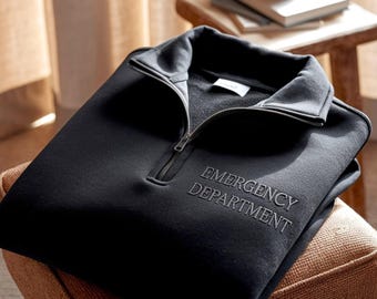 Puede incluir: Sudadera negra con cremallera de un cuarto de cuello con las palabras "EMERGENCY DEPARTMENT" bordadas en la parte delantera. La sudadera tiene un cuello alto y una cremallera plateada. La prenda está doblada y apoyada en un pequeño taburete tapizado.