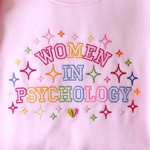 May include: A light pink sweatshirt featuring multi-colored embroidered text that reads WOMEN IN PSYCHOLOGY. The design is surrounded by various small, colorful embroidered star shapes and a single small heart icon at the bottom center.