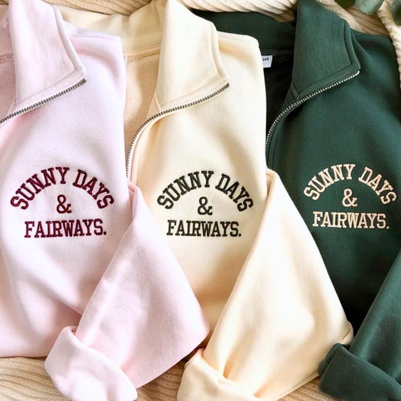 Puede incluir: Primer plano de tres sudaderas en rosa pastel, crema y verde oscuro. Cada una tiene un cuello con cremallera y el texto bordado "SUNNY DAYS & FAIRWAYS". Las mangas est&aacute;n dobladas y la tela parece suave y c&oacute;moda.