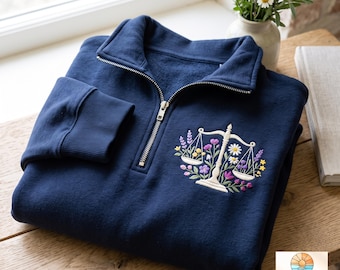 May include: A navy blue quarter-zip sweatshirt featuring an embroidered design of a white scale of justice surrounded by colorful flowers including daisies, lavender, and small yellow blossoms. The sweatshirt has a silver metal zipper and ribbed cuffs.