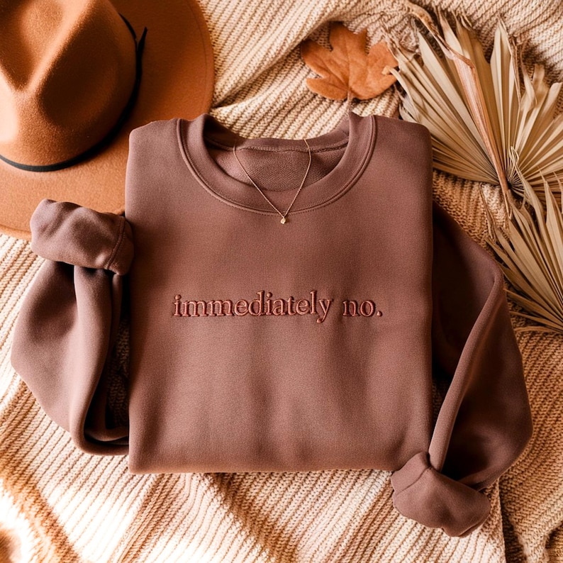 Peut inclure: Un sweat-shirt marron &agrave; col rond avec le texte brod&eacute; "immediately no." Un collier en or avec un petit pendentif est drap&eacute; sur le sweat-shirt. Un chapeau marron et des plantes s&eacute;ch&eacute;es d&eacute;coratives sont en arri&egrave;re-plan.