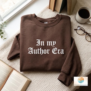 Puede incluir: Una sudadera de cuello redondo en color marrón oscuro con el texto blanco In my Author Era en una fuente de estilo gótico. La prenda tiene mangas largas y cuello, puños y dobladillo acanalados, ofreciendo un aspecto sencillo y limpio.