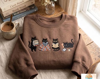May include: A brown crewneck sweatshirt featuring an embroidered graphic of four black cats engaged in reading activities. One cat holds a stack of books, another is wrapped in a blanket, one wears glasses while reading, and the last lies by a tablet.