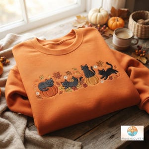 May include: An orange crewneck sweatshirt featuring an embroidered design of three black cats playing among autumn leaves, small white flowers, and orange pumpkins. The embroidery is centred on the chest of the garment.