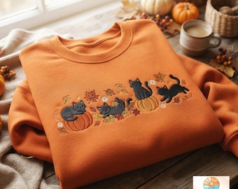 Op de afbeelding: Een oranje sweater met ronde hals voorzien van een borduursel van drie zwarte katten die spelen tussen herfstbladeren, kleine witte bloemen en oranje pompoenen. Het borduurwerk is gecentreerd op de borst van het kledingstuk.