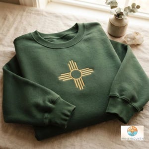 May include: A forest green crewneck sweatshirt featuring a gold embroidered Zia sun symbol centered on the chest. The garment has long sleeves with ribbed cuffs and a ribbed neckline, showcasing a clean, minimalist design with a soft fabric texture.