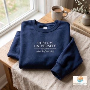 Puede incluir: Una sudadera azul marino de cuello redondo con texto blanco que dice CUSTOM UNIVERSITY EST. 1972 school of nursing. La prenda tiene mangas largas, un cuello acanalado y un interior de forro polar suave, presentada como una prenda doblada.