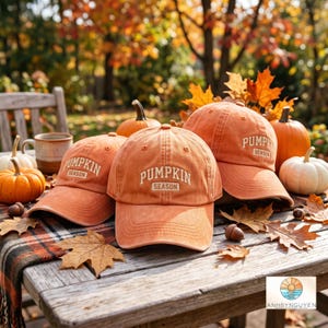 May include: Three orange baseball caps featuring the text PUMPKIN SEASON embroidered in white on the front panels. The caps have a structured crown, a curved brim, and eyelets for ventilation, displayed alongside pumpkins and autumn leaves.