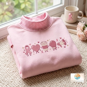 Op de afbeelding: Een lichtroze sweatshirt met een opstaande kraag in een roze en wit gingham-patroon. Op de borst staan geborduurde cartoon-iconen, waaronder een lint voor borstkankerbewustzijn, een hart, een koffiebeker, een bokshandschoen en een megafoon.