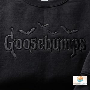May include: A black sweatshirt featuring the word Goosebumps embroidered in a matching black thread across the chest. Above the text, three small black bats are embroidered in a row. The fabric has a visible texture and a matte finish.