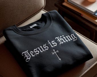 May include: A black crewneck sweatshirt featuring white embroidered text that reads "Jesus is King" in a gothic-style font, positioned above a small, simple embroidered white cross centered on the chest area of the garment.