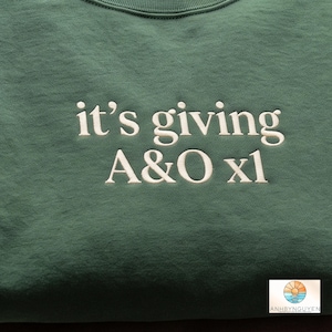 Puede incluir: Una sudadera verde bosque con texto bordado en blanco que dice "it's giving A&O xl" centrado en el pecho. La prenda tiene un cuello redondo acanalado y un acabado de tela suave y texturizada, mostrando costuras claras y precisas.