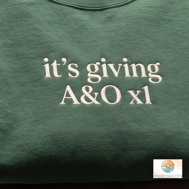 Puede incluir: Una sudadera verde bosque con texto bordado en blanco que dice "it's giving A&O xl" centrado en el pecho. La prenda tiene un cuello redondo acanalado y un acabado de tela suave y texturizada, mostrando costuras claras y precisas.