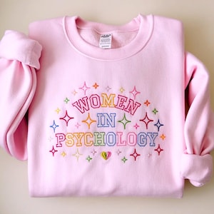 May include: A light pink crewneck sweatshirt featuring embroidered text that reads "WOMEN IN PSYCHOLOGY" in a multi-colored block font. The design is surrounded by various small, colorful embroidered starburst shapes and a small heart at the bottom.