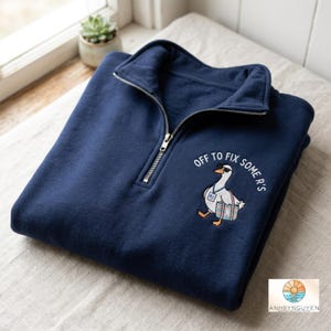 May include: A navy blue quarter-zip sweatshirt featuring an embroidered graphic of a white goose wearing sunglasses, a lanyard, and carrying a striped bag. The text above the goose reads "OFF TO FIX SOME R'S" in white, capitalised, arched lettering.
