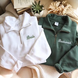 May include: Two sweatshirts with a quarter-zip collar. One is white with "Bride" embroidered in green, the other is dark green with "Bridesmaid" embroidered in white. Both have long sleeves and a relaxed fit, perfect for a bridal party.