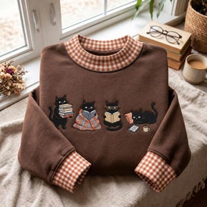May include: A brown long-sleeve sweatshirt featuring a gingham collar and cuffs. The front displays an embroidered graphic of four black cats engaged in reading activities, including holding books, wearing glasses, and resting near a tablet.