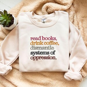 May include: A light pink sweatshirt with the embroidered text "read books, drink coffee, dismantle systems of oppression." The sweatshirt is made of a soft material and is displayed on a fluffy, beige surface.