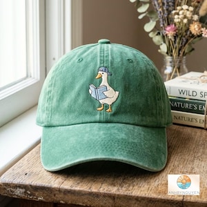 Op de afbeelding: Een bosgroene baseballpet met een geborduurde afbeelding van een witte gans die een blauwe pet draagt en een lichtblauw boek leest. De pet heeft een gestructureerde kroon, een gebogen klep en zichtbare stiksels op de voorste panelen.