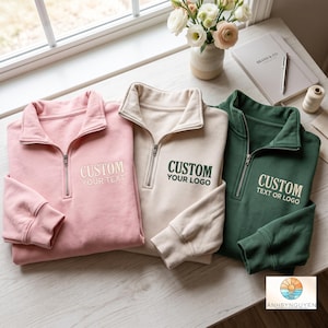 Peut inclure: Trois sweat-shirts à fermeture éclair quart de longueur en rose clair, beige et vert foncé. Chacun présente un texte brodé sur la poitrine gauche : le rose indique CUSTOM YOUR TEXT, le beige CUSTOM YOUR LOGO et le vert CUSTOM TEXT OR LOGO.