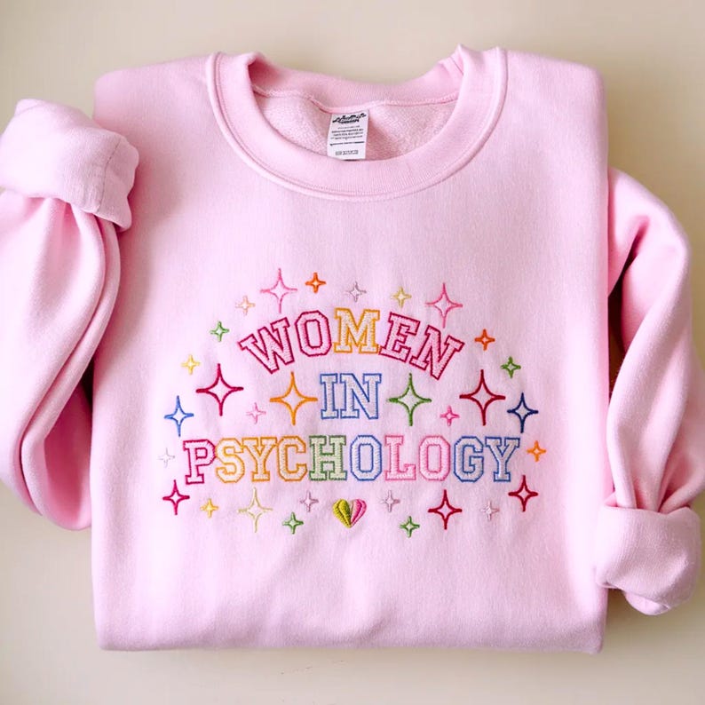 May include: A light pink crewneck sweatshirt featuring embroidered text that reads "WOMEN IN PSYCHOLOGY" in a multi-colored block font. The design is surrounded by various small, colorful embroidered starburst shapes and a small heart at the bottom.