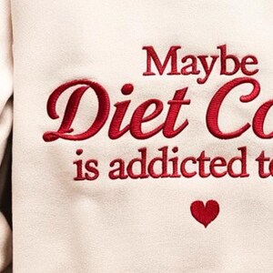 Może przedstawiać: Kremowa bluza z czerwonym haftowanym napisem: "Maybe Diet Coke is addicted to me". Małe czerwone serce znajduje się pod tekstem. Grafika puszki Diet Coke znajduje się na rękawie.