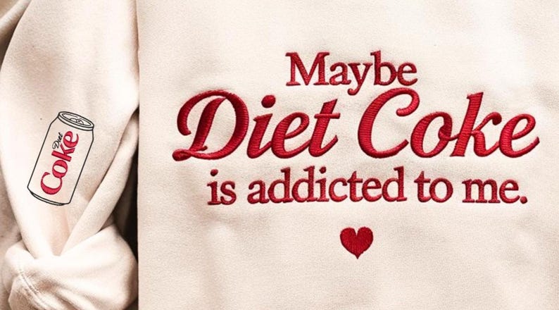 Może przedstawiać: Kremowa bluza z czerwonym haftowanym napisem: "Maybe Diet Coke is addicted to me". Małe czerwone serce znajduje się pod tekstem. Grafika puszki Diet Coke znajduje się na rękawie.