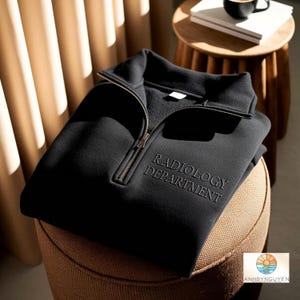 Puede incluir: Una sudadera negra con cremallera de un cuarto, cuello alto y cremallera de metal plateado. El texto RADIOLOGY DEPARTMENT está bordado con hilo negro a juego en el lado derecho del pecho, creando un sutil efecto de textura tono sobre tono.
