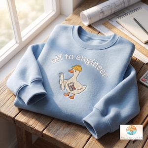 Könnte beinhalten: Ein hellblaues Sweatshirt mit Rundhalsausschnitt und einer Stickerei, die eine weiße Gans mit gelbem Schutzhelm zeigt, welche einen Bauplan und einen Taschenrechner hält, sowie dem Schriftzug "off to engineer" in weißen Buchstaben.