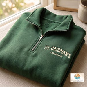 Puede incluir: Una sudadera verde bosque con cremallera de un cuarto, cuello alto y cremallera de metal plateado. El pecho muestra un texto bordado en color crema que dice ST. CRISPIAN'S LONDON en una fuente sans-serif limpia.